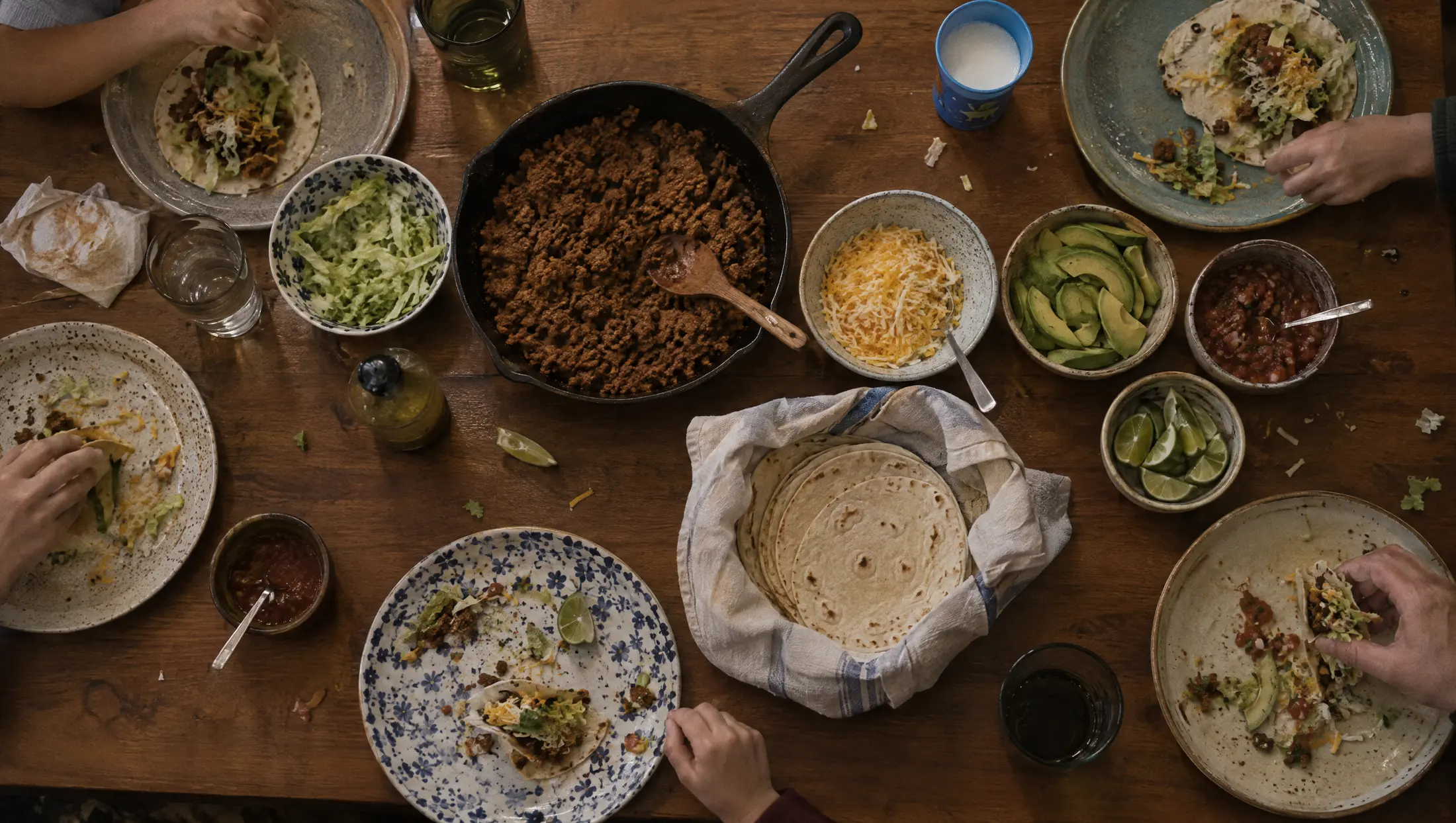 Taco night seen from above: a pan of meat, bowls of cheese, avocado and limes, tortillas in a towel, hands reaching in.